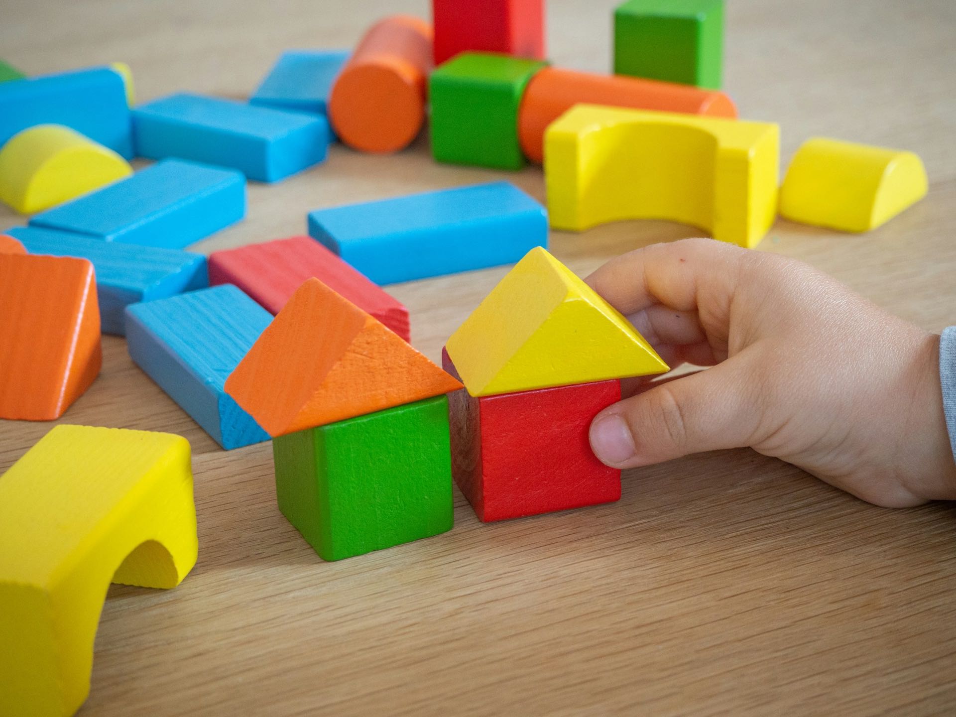 a child's hand fitting a triangle block onto a small wooden block house, colorful building blocks scattered on a table